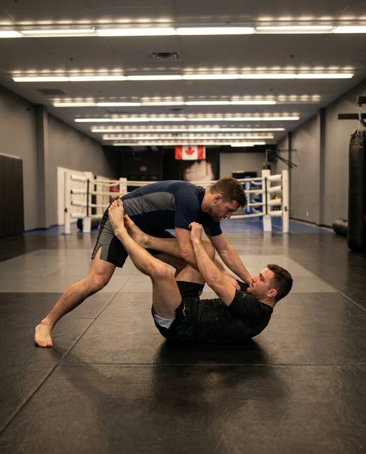 Two adults drilling a grappling technique on the mats at Rumble Martial Arts, with the white boxing ring behind them.