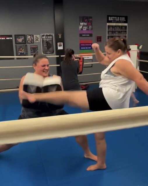 A Battle Girl member landing a round kick into Thai pads held by her partner, inside the ring on the blue mats.