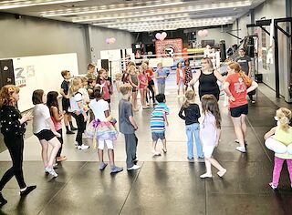 A circle of children playing a party game on the black mats at Rumble Martial Arts, led by an instructor, with the boxing ring behind them.