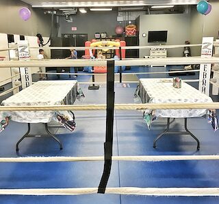 Two party tables with tablecloths set up on the blue mats beside the boxing ring, with balloons and an inflatable play area behind.