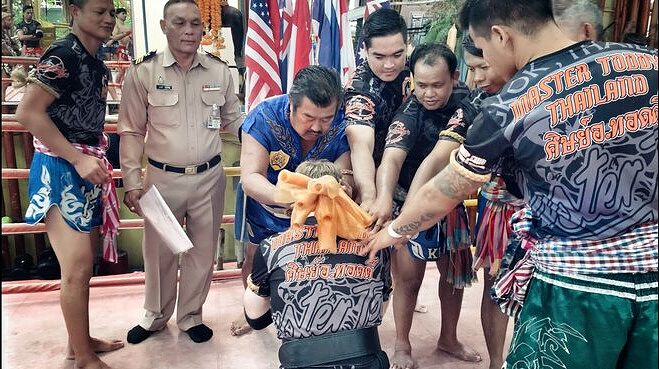 Kru certification ceremony in Thailand: Master Toddy and his instructors gathered around Kru Jones, kneeling, placing the Mongkon on Kru Jones's head.