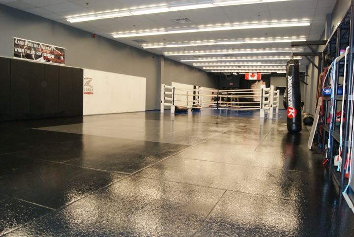 The main mat floor at Rumble Martial Arts: black mats, a white boxing ring, a heavy bag and a Canadian flag.