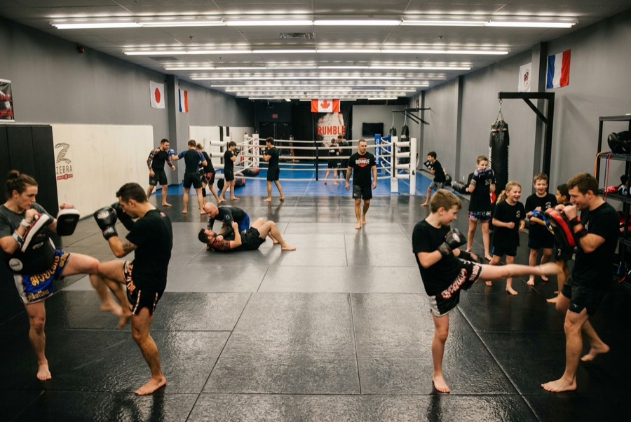 A busy evening at Rumble Martial Arts: adults working Thai pads in the foreground, adults drilling jiu-jitsu on the left, kids doing pad work with a coach on the right, and the boxing ring at the back under the Canadian flag.