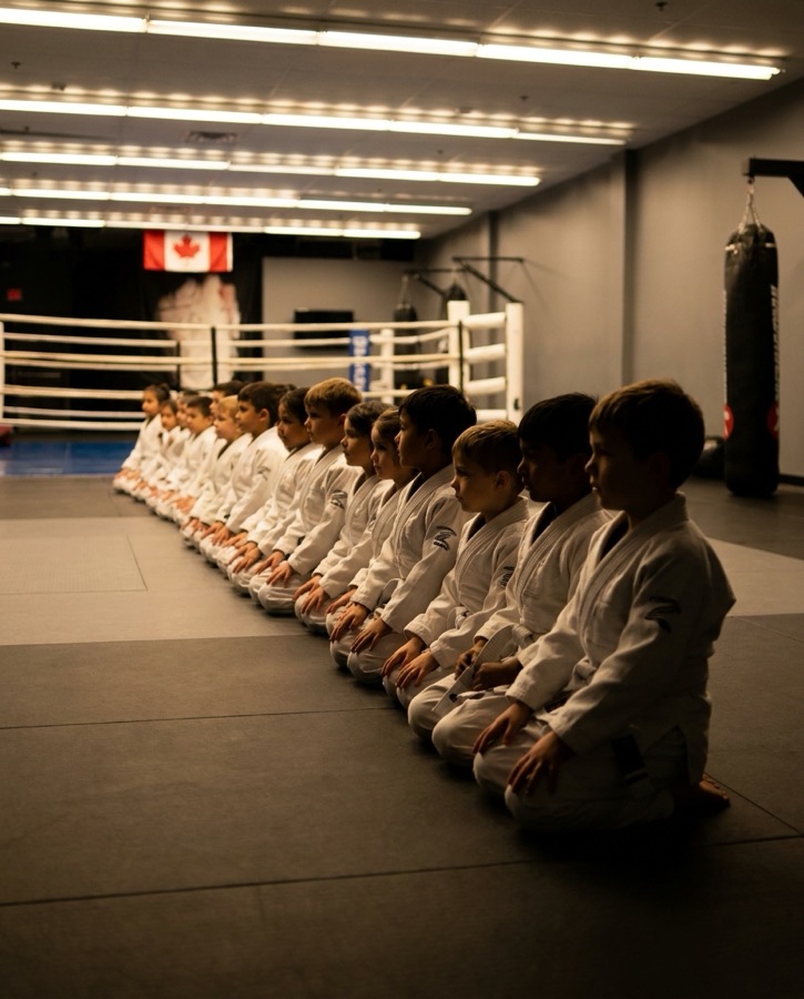 Young children in white Brazilian Jiu-Jitsu Gis kneeling on the mats at Rumble Martial Arts in Keswick, listening to their coach