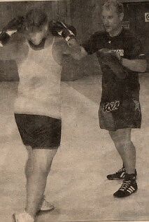 Grainy black-and-white newspaper photo of Kru Jones, in a dark t-shirt and shorts, holding focus pads for a person in a white tank top and dark shorts who has gloves raised mid-strike, on the floor of a hall.