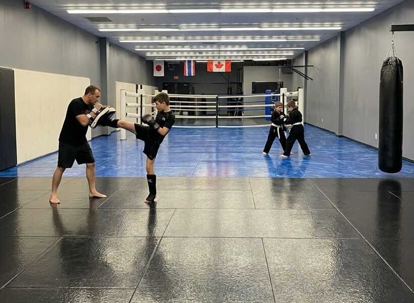 A teen landing a kick on Thai pads held by a coach at Rumble Martial Arts, with younger kids training nearby.