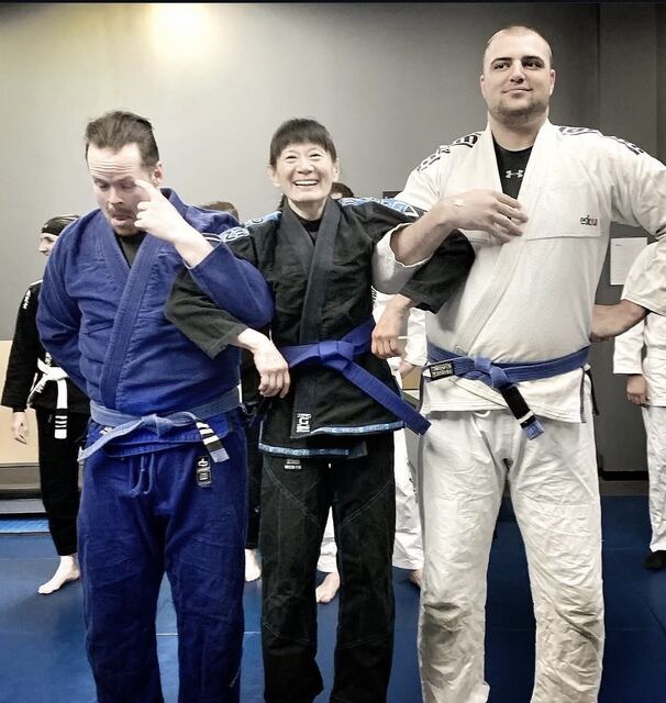 Coach Gladys on the mats at Rumble after a belt promotion, standing between two teammates.