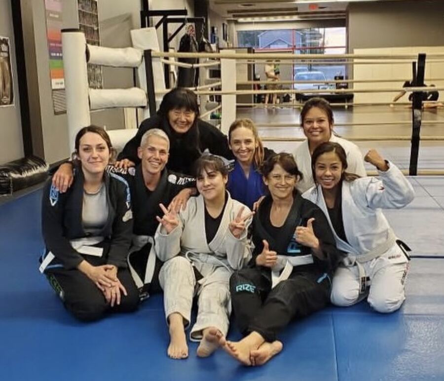 The women's BJJ group in their gis on the blue mats at Rumble, smiling with Coach Gladys.